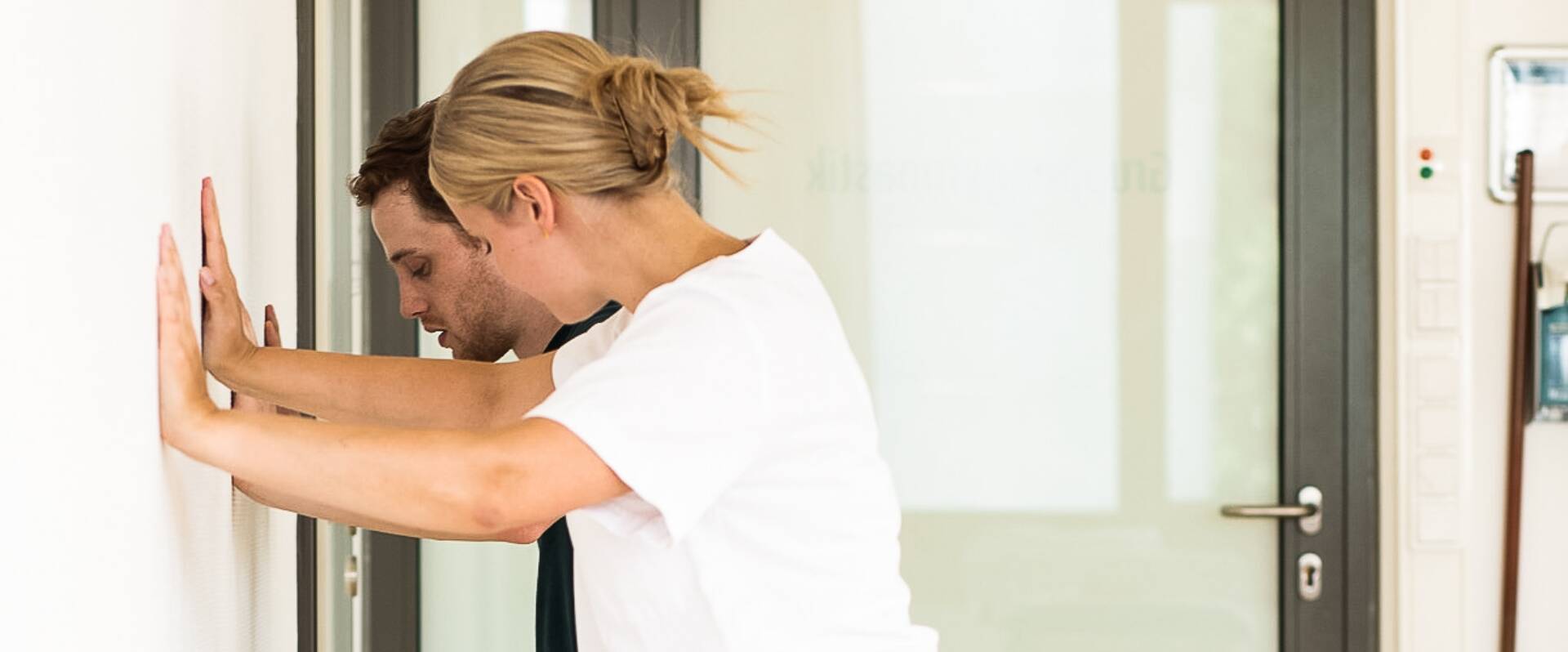 Physiotherapeutin zeigt Kraftübung an der Wand mit einem Patienten