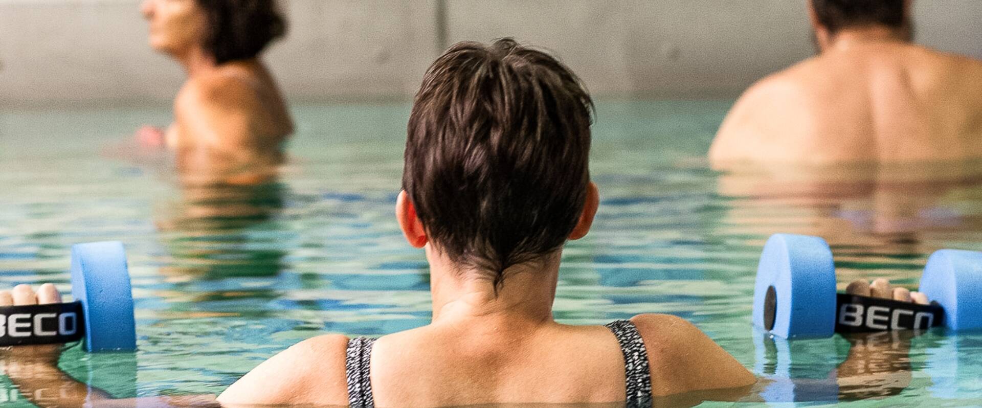 Patientin von hinten im Schwimmbecken mit zwei Hanteln 